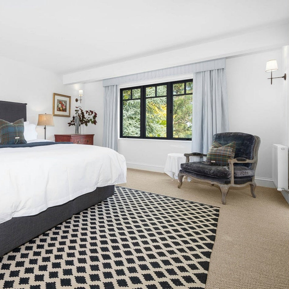 Sunlit bedroom featuring a bed with plaid pillow, armchair, blue curtains, and a fine rustic sisal rug on timber floors.