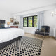Sunlit bedroom featuring a bed with plaid pillow, armchair, blue curtains, and a fine rustic sisal rug on timber floors.