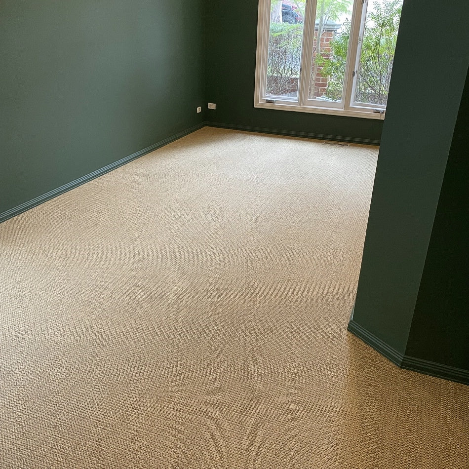 A serene space with green walls, beige carpet, and a handwoven sisal rug in subtle autumnal tones placed by a sunlit window.