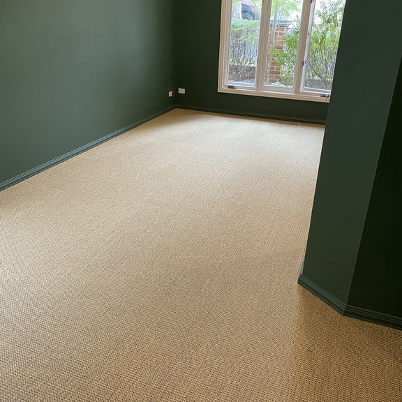 A serene space with green walls, beige carpet, and a handwoven sisal rug in subtle autumnal tones placed by a sunlit window.