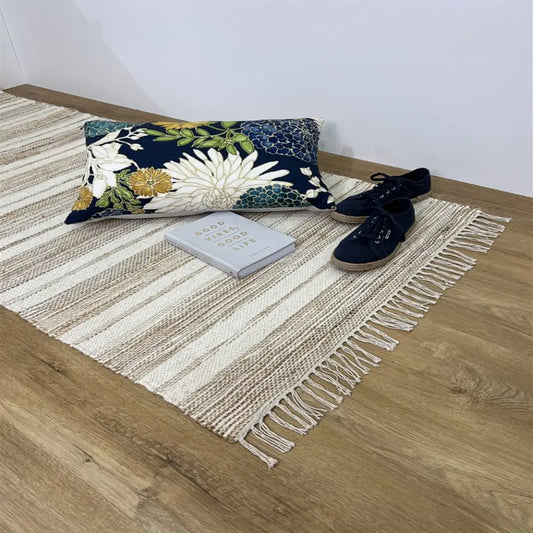 A handwoven natural fibre rug lies on warm timber flooring, styled with a floral cushion, open book and black flats.