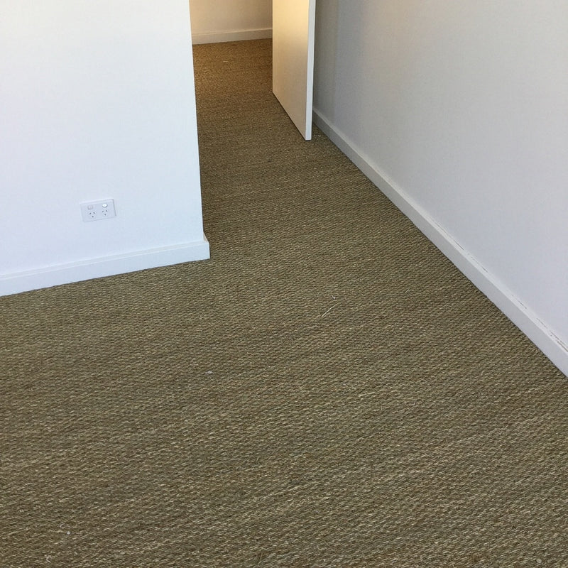 A minimalist room with white walls, beige carpet, and an open door revealing elegant basketweave seagrass flooring beyond.