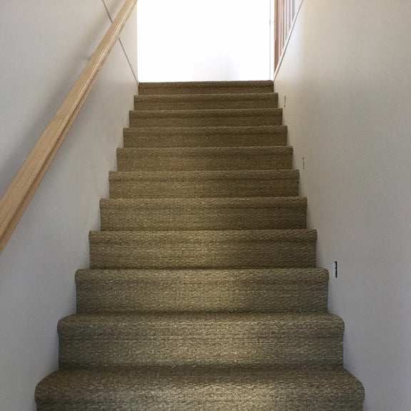 A staircase clad in woven seagrass carpet with a timber handrail leads to a sunlit landing in a refined, contemporary home.