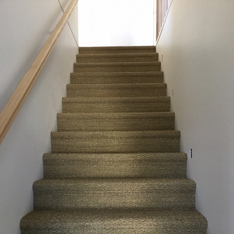 A staircase clad in woven seagrass carpet with a timber handrail leads to a sunlit landing in a refined, contemporary home.