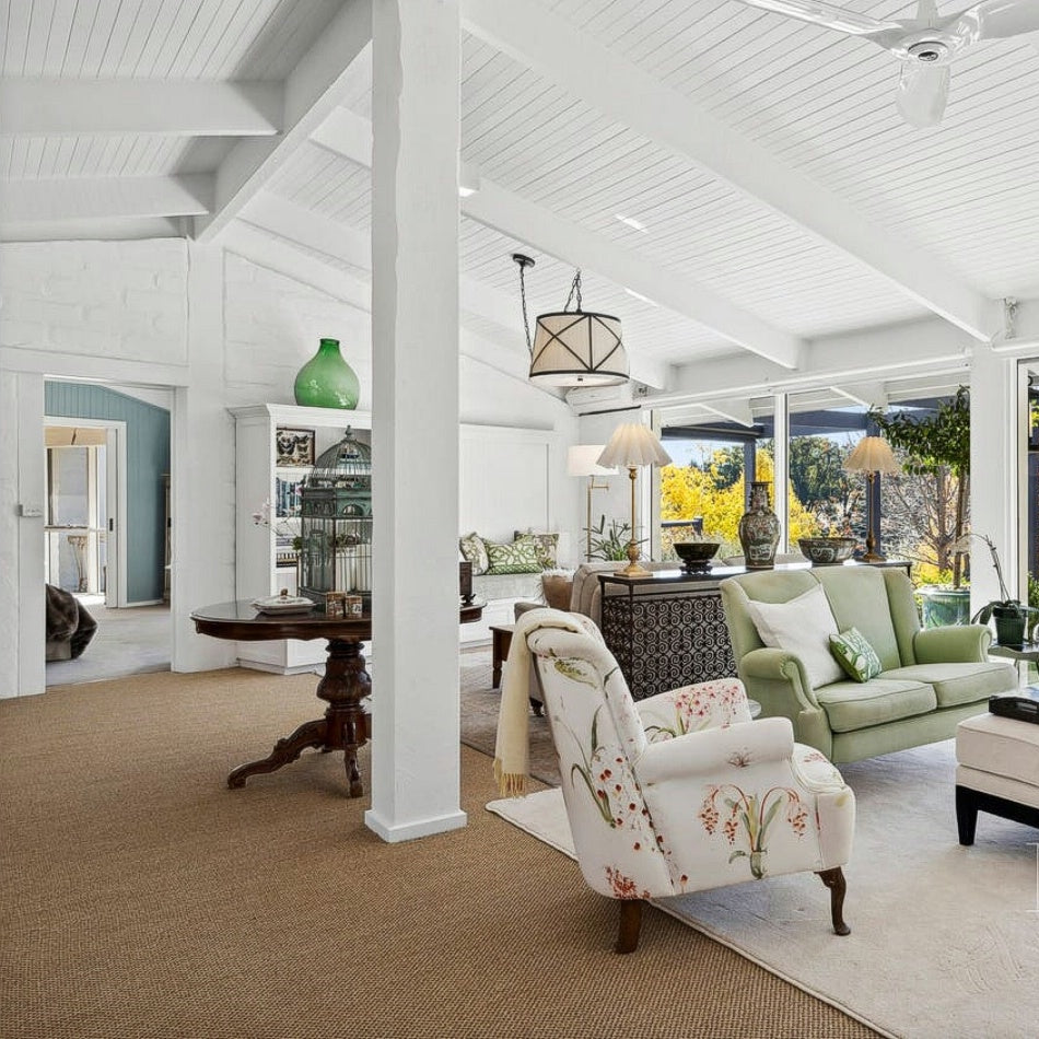 Sunlit living room with white walls, floral armchair, green sofa, and a textured sisal rug anchoring the elegant space.