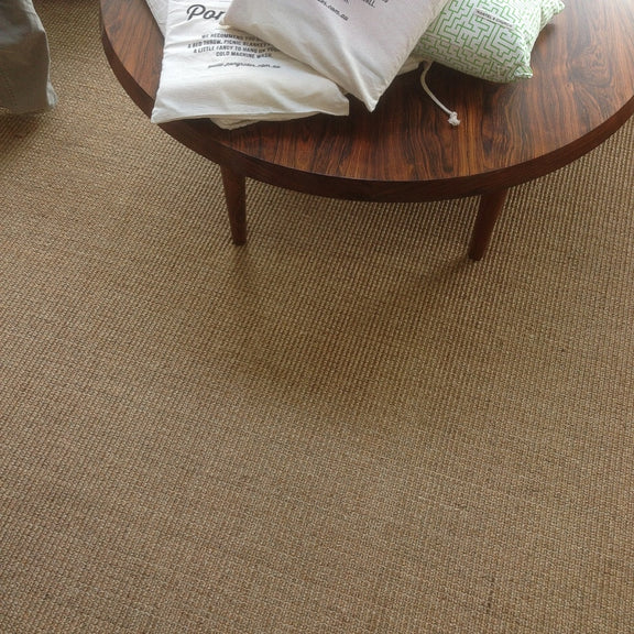 A round wooden coffee table with tote bags and a pillow rests on richly textured, natural sisal flooring in a styled interior.