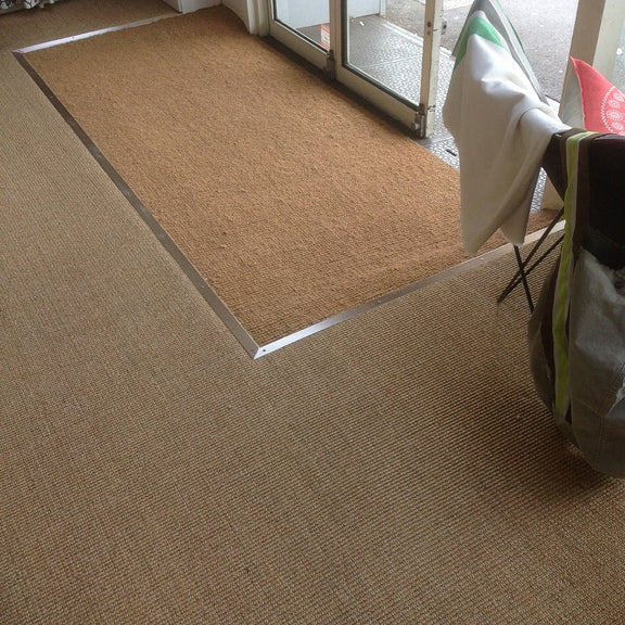 A brown doormat and shopping bags rest at a glass door, set atop textured, natural sisal flooring in a refined entryway.