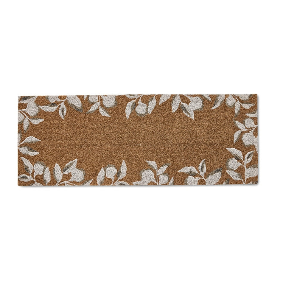Rectangular natural fibre doormat, warm brown tone with refined white leafy branch edging; styled on a modern entryway floor.