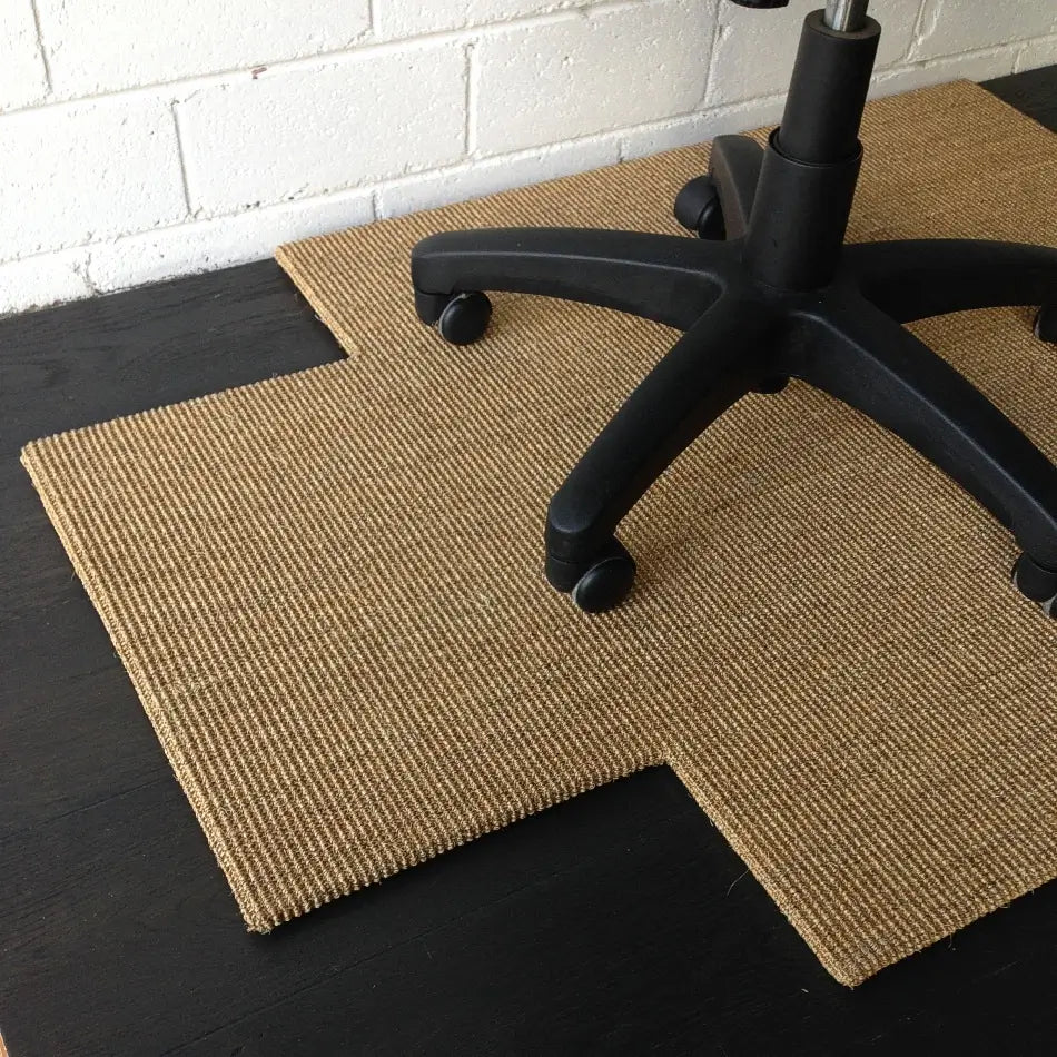 A tan, cross-shaped sisal chair pad sits beneath a black office chair on hardwood flooring in a refined, modern workspace.