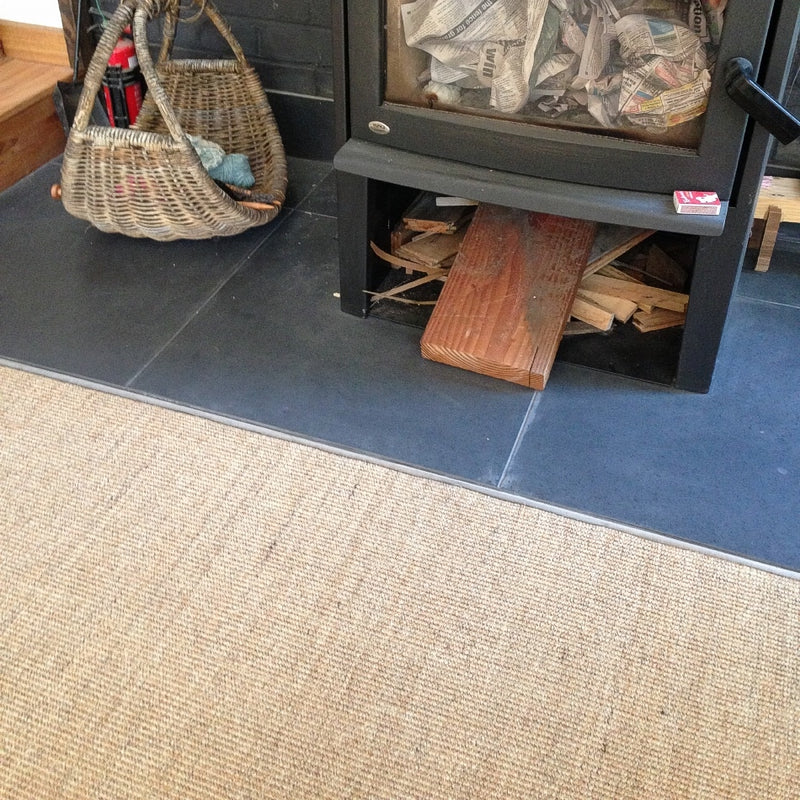 A wood stove on a tiled hearth, with kindling and papers resting on a finely woven sisal rug in a thoughtfully styled living space.