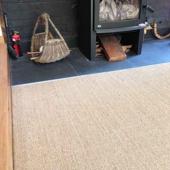 A fine ribbed sisal rug lies beside a modern fireplace with neatly stacked firewood, woven basket, and extinguisher on black tiles.