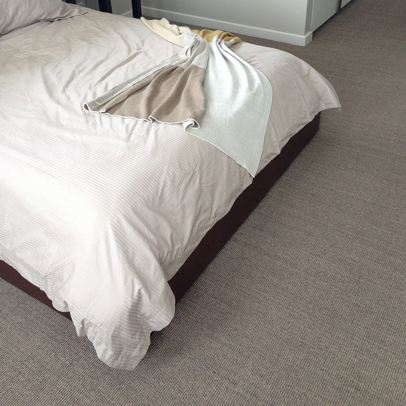 An unmade bed with soft, light bedding and a neatly folded blanket rests on fine ribbed granite sisal flooring in a serene interior.
