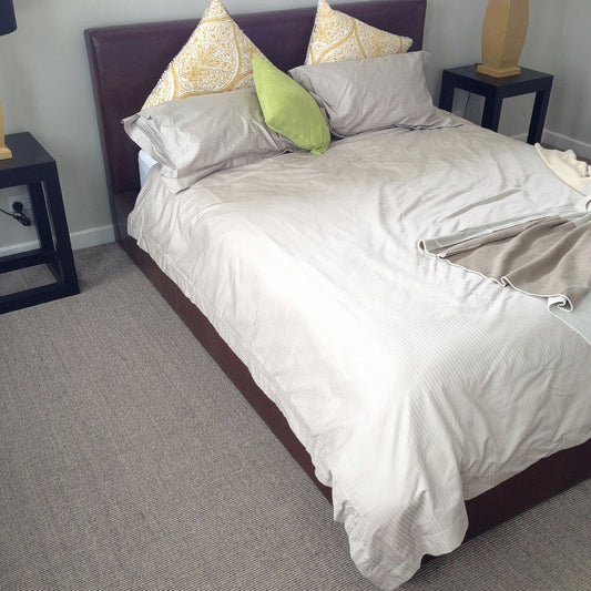 A serene bedroom with a beige duvet, colourful accent pillows, black side tables and a textured granite sisal rug underfoot.