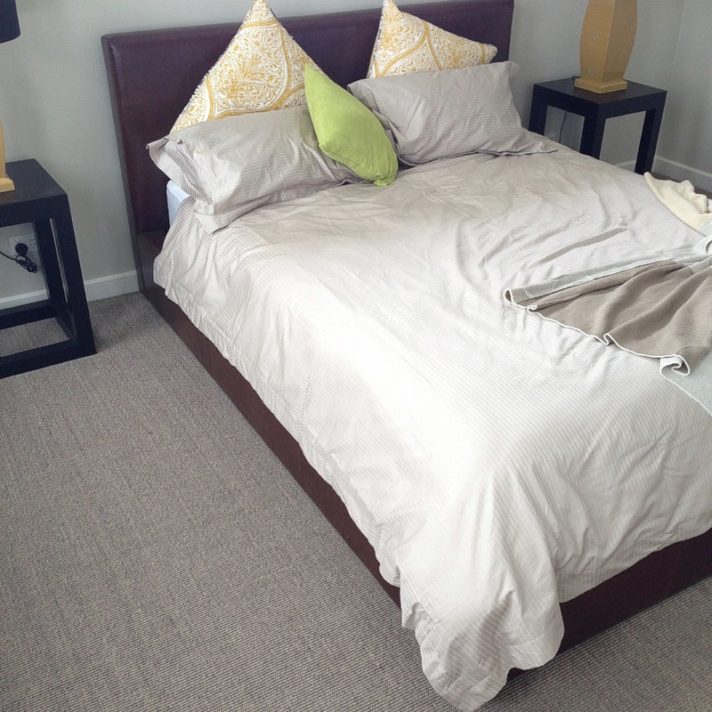 A serene bedroom with a beige duvet, colourful accent pillows, black side tables and a textured granite sisal rug underfoot.