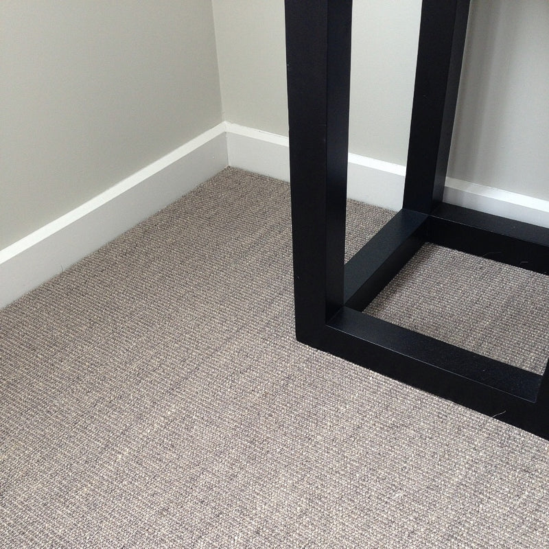 A sleek black table leg rests on fine ribbed granite sisal carpet beside crisp white baseboards and pale grey walls.