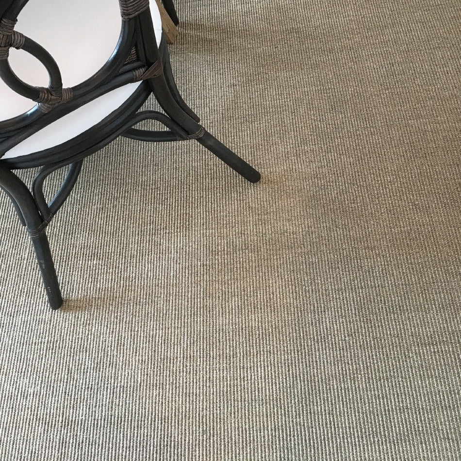 A black wicker chair with a white cushion sits on fine ribbed quartz sisal flooring, styled in an elegant, modern interior.