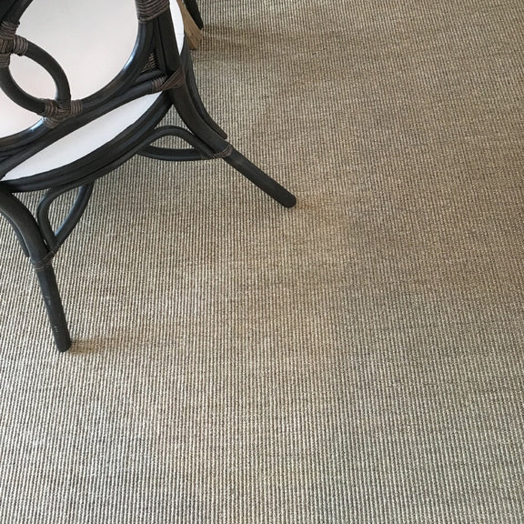 A black wicker chair with a white cushion sits on fine ribbed quartz sisal flooring, styled in an elegant, modern interior.