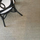 A black wicker chair with a white cushion sits on fine ribbed quartz sisal flooring, styled in an elegant, modern interior.