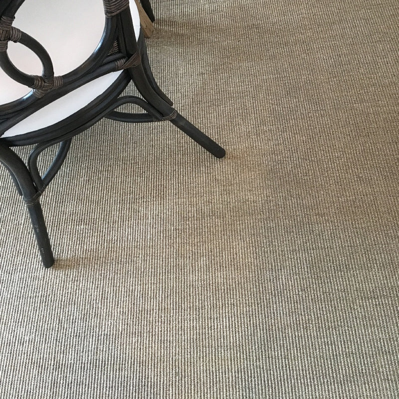 A black wicker chair with a white cushion sits on fine ribbed quartz sisal flooring, styled in an elegant, modern interior.
