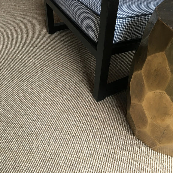 A checkered cushion chair sits on fine ribbed quartz sisal, beside a gold geometric side table in a thoughtfully styled interior.