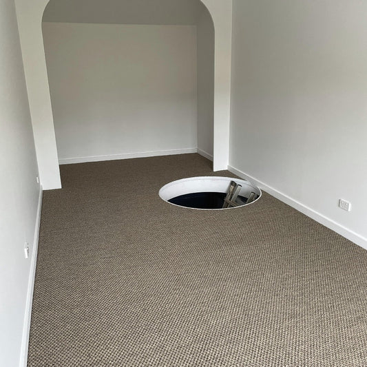 A contemporary room with fine rustic sisal carpet, highlighted by a floor opening and sleek ladder descending below.