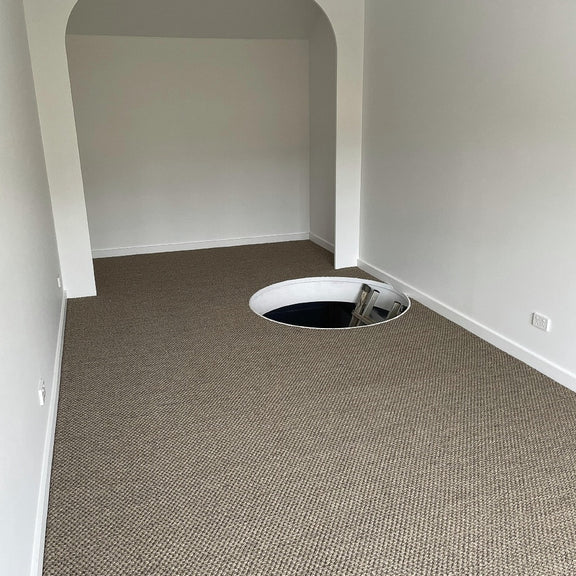 A contemporary room with fine rustic sisal carpet, highlighted by a floor opening and sleek ladder descending below.