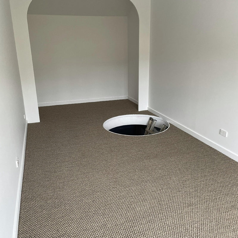 A contemporary room with fine rustic sisal carpet, highlighted by a floor opening and sleek ladder descending below.