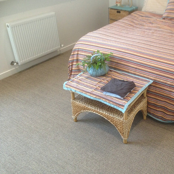A small wicker table with a potted plant and neatly folded clothes sits beside a bed on natural sisal flooring in a serene bedroom.