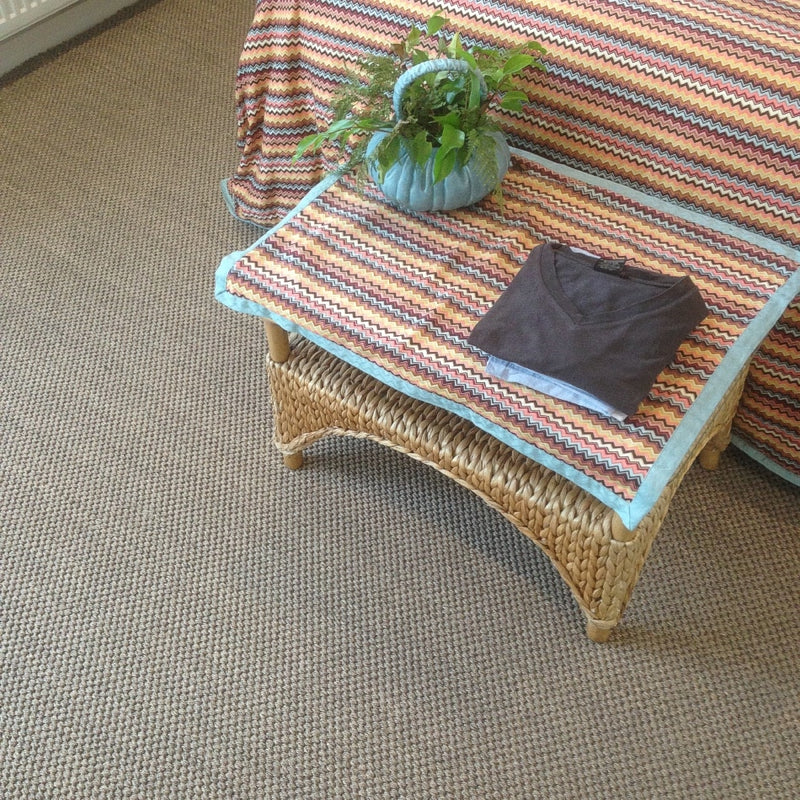 A handwoven wicker table holds a folded shirt and leafy plant beside a bed on textured granite-toned sisal flooring.