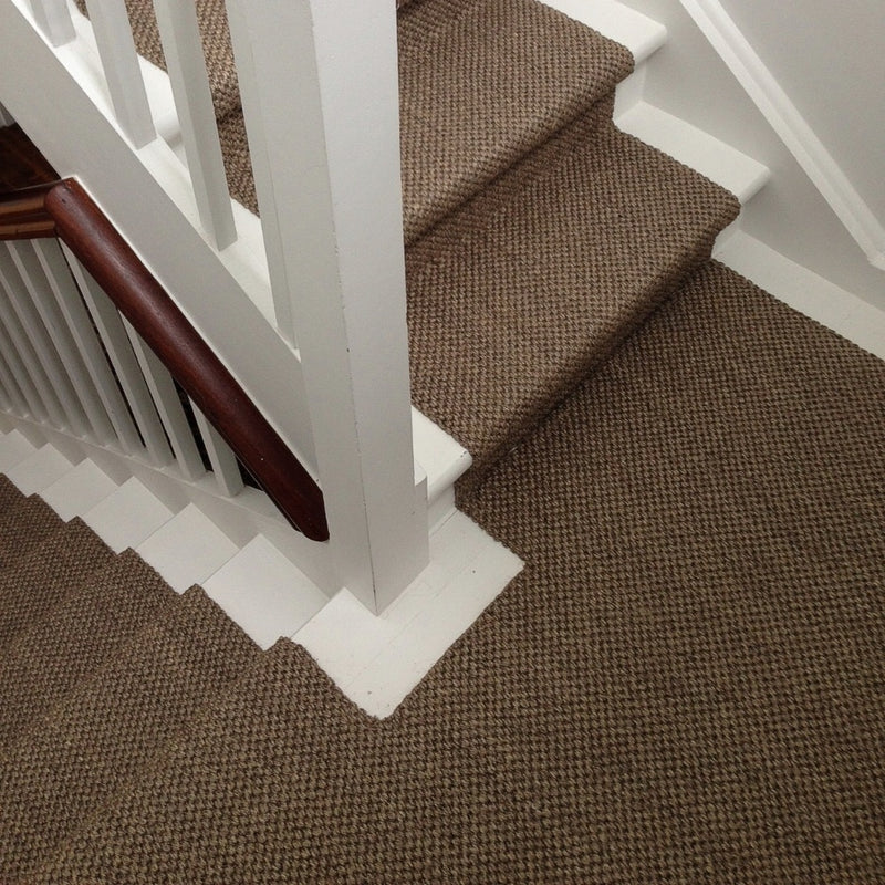 A staircase clad in textured, natural sisal carpet with crisp white trim and a smooth timber handrail, viewed from above.