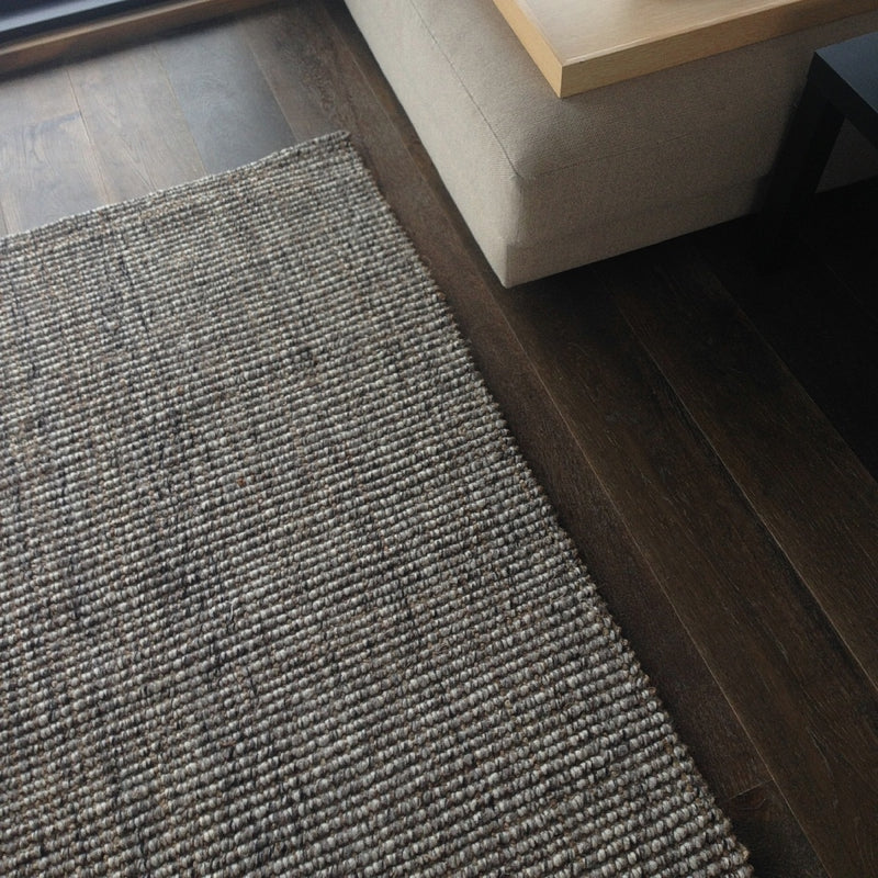 A greystone wool and jute rug rests on rich dark wood flooring next to a beige sofa and sleek black side table in a styled interior.