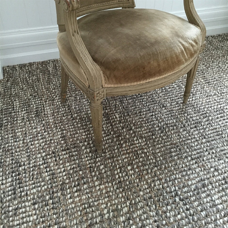 A wooden armchair with tan velvet upholstery sits on a textured greystone wool and jute rug, set against a crisp white wall.