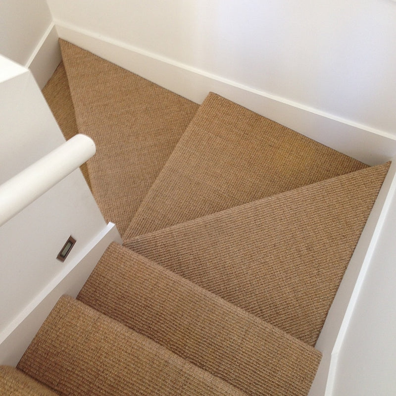 Sisal carpet with a refined heavy ribbed texture lines a corner staircase with crisp white walls and minimalist handrail.