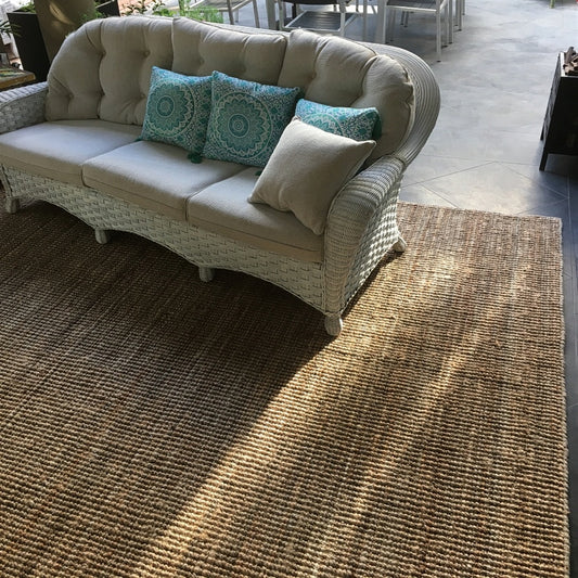 A wicker sofa with plush blue and neutral cushions rests on a natural jute rug, paired with a dining table in a serene living space.