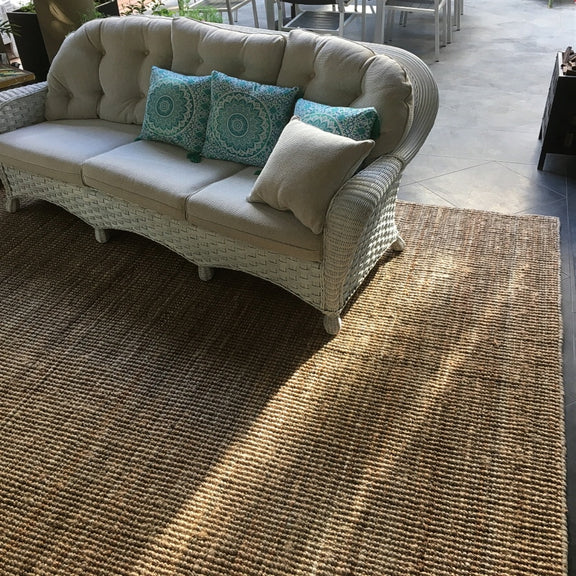 A wicker sofa with plush blue and neutral cushions rests on a natural jute rug, paired with a dining table in a serene living space.