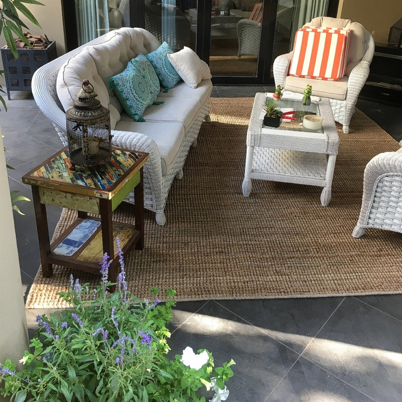 Wicker patio chairs with plush cushions arranged on a natural jute rug, complemented by side tables and potted flowers.