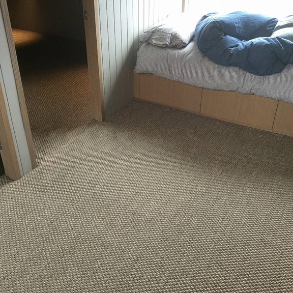 Blue bedding on an unmade bed next to a rustic granite-hued sisal rug in a softly lit, tastefully styled bedroom.
