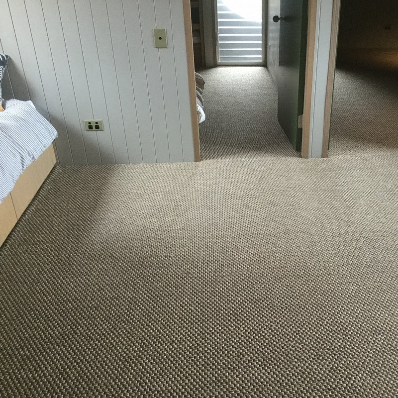 A serene bedroom with textured granite-coloured sisal flooring, a neatly made bed and an open doorway leading to a hallway.