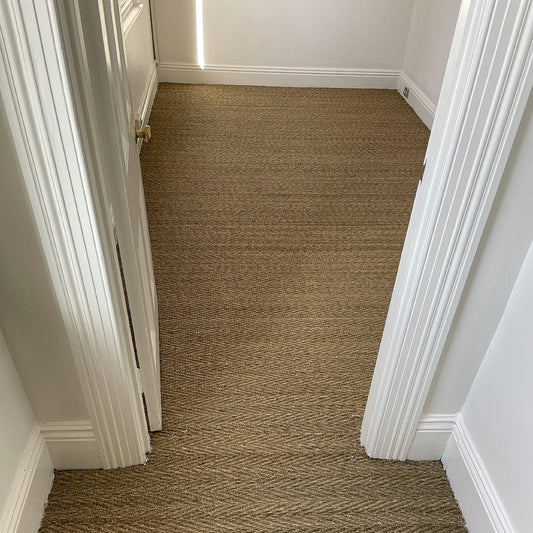 A bright, small room with crisp white walls and elegant herringbone seagrass flooring viewed through an open doorway.