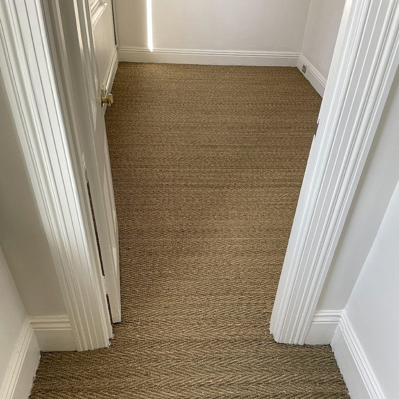 A bright, small room with crisp white walls and elegant herringbone seagrass flooring viewed through an open doorway.