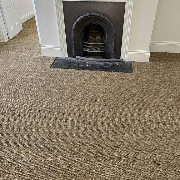 Elegant herringbone seagrass flooring highlights a modern fireplace in a bright, white-walled, thoughtfully styled living space.