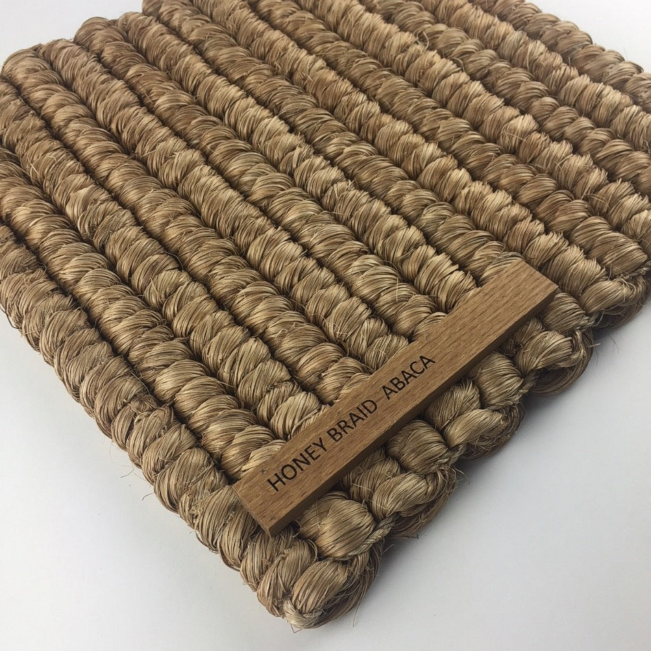 A richly textured honey-toned mat crafted from thick, braided abaca fibers with a subtle wooden label at one corner.