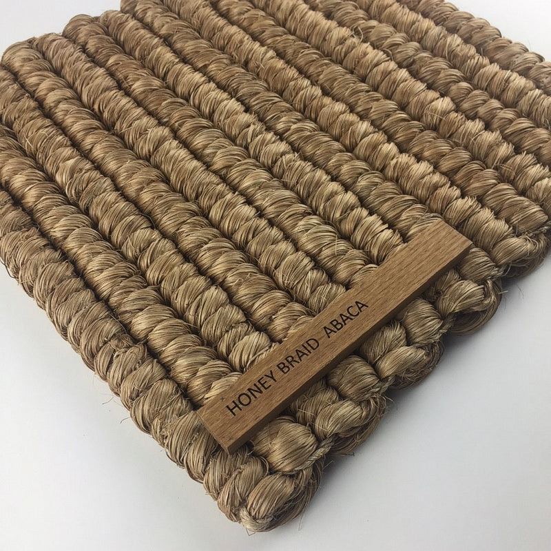 A richly textured honey-toned mat crafted from thick, braided abaca fibers with a subtle wooden label at one corner.