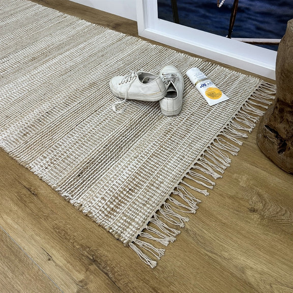 A pair of white sneakers and sunscreen rest on a textured natural fibre runner in a sunlit, tastefully styled entryway hallway.