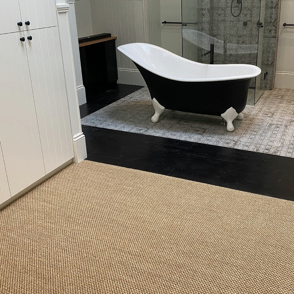 A black and white clawfoot tub stands on textured quartz-toned sisal flooring in an elegantly styled, light-filled bathroom.