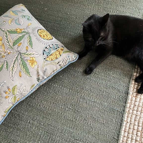 A sleek black cat lounges on an elegant moss-toned jute rug, beside a botanical print pillow in a softly styled living space.