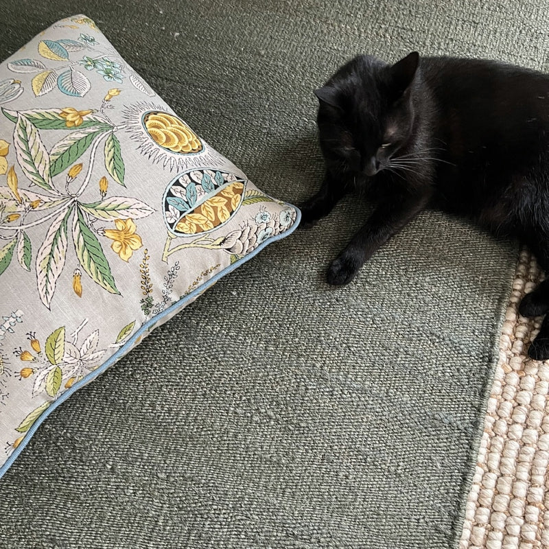 A sleek black cat lounges on an elegant moss-toned jute rug, beside a botanical print pillow in a softly styled living space.