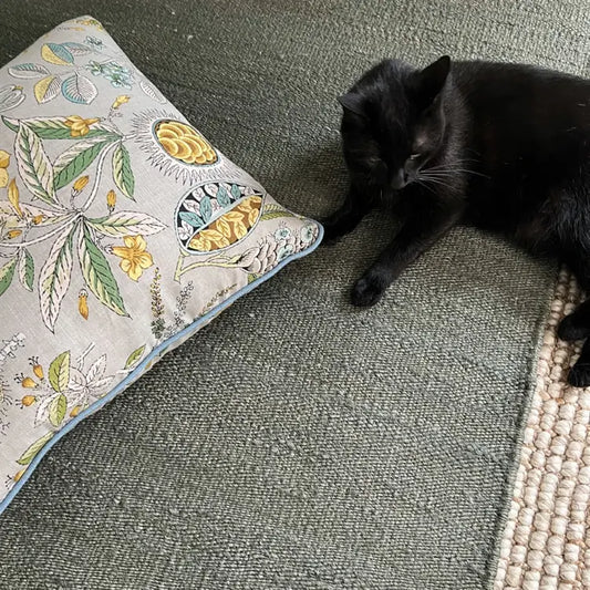A sleek black cat lounges on a textured moss-green jute rug, beside a floral and lemon print cushion in an elegant living space.