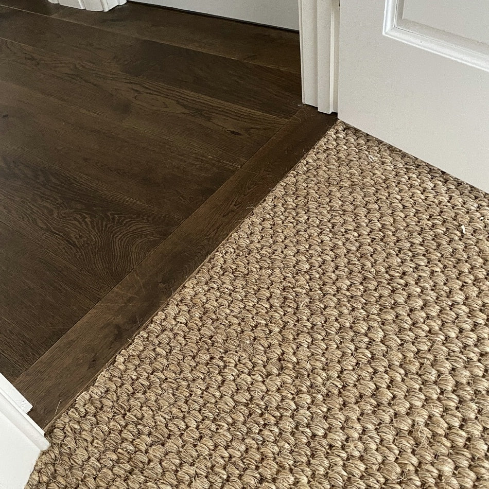 Rich dark hardwood flooring seamlessly transitions to a textured sisal rug in soft river rock tones, styled beside a white doorway.
