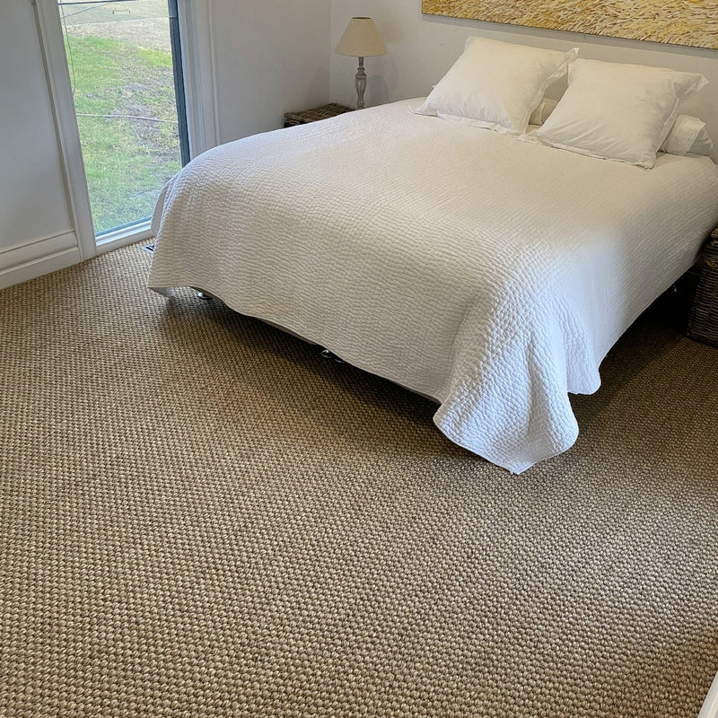 A bright room features a plush white bed atop a textured, handwoven sisal rug in a refined rustic river rock hue.